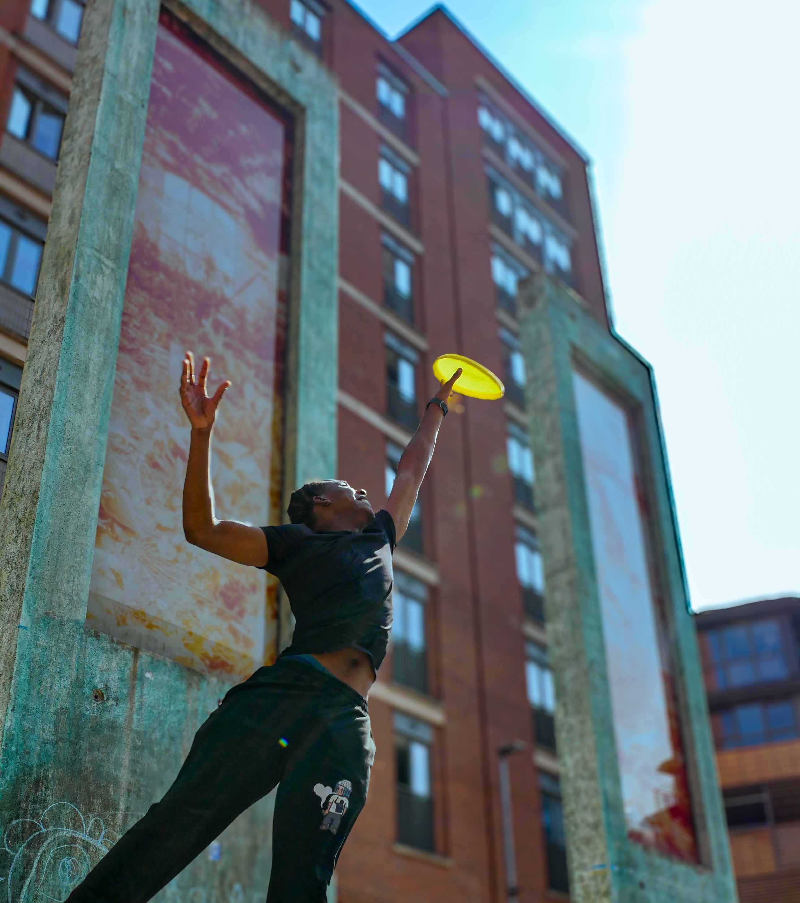 A person in black athletic wear jumps to catch a yellow frisbee outdoors in front of a brick building with large painted panels.